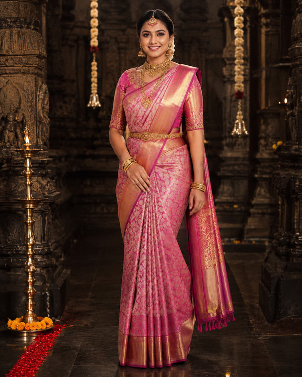 Woman in a pink saree with gold details standing in an ornate setting.